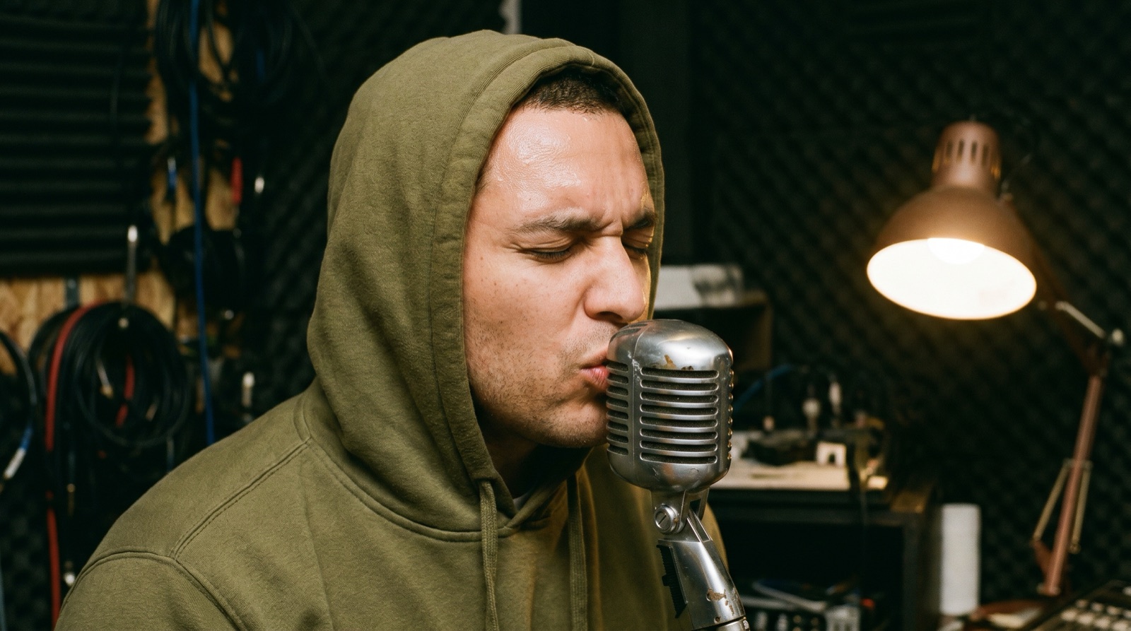 Un Beatboxer con sudadera color oliva actuando muy cerca de un micrófono vintage en un estudio casero