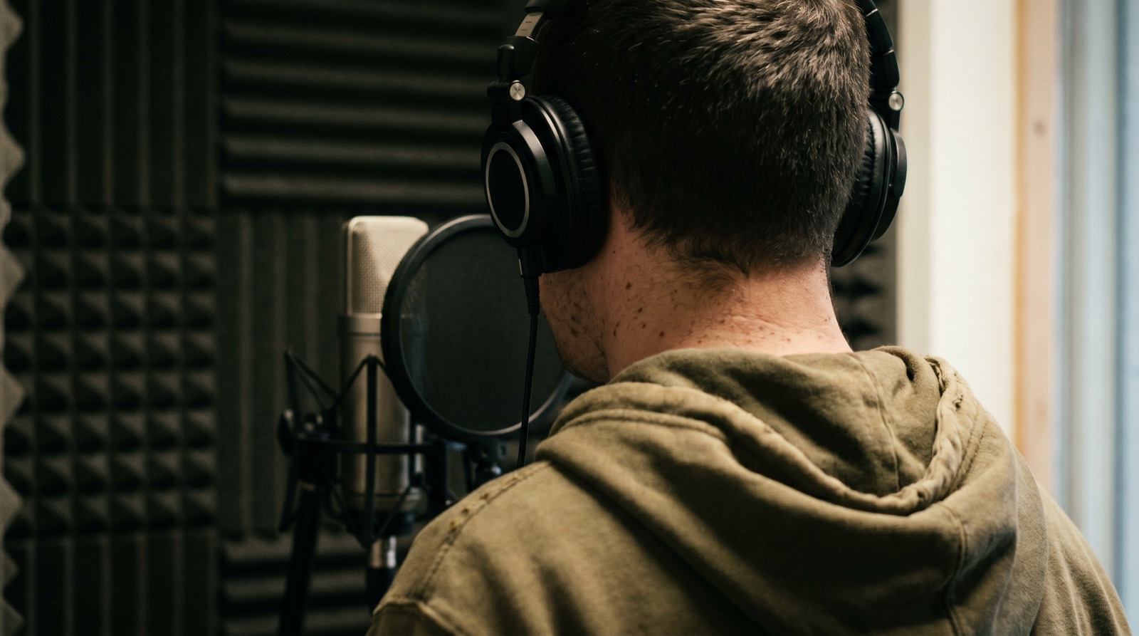 Un Beatboxer con auriculares grabando en un micrófono de condensador de estudio dentro de una cabina insonorizada, visto desde atrás