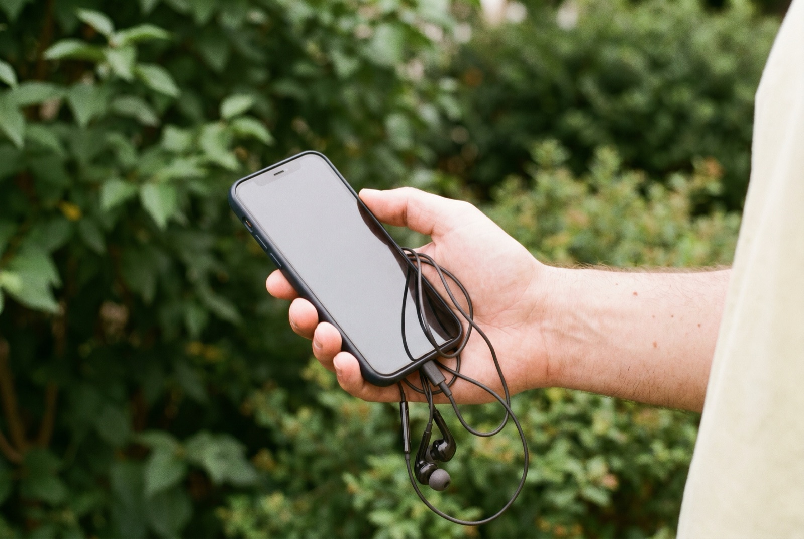 Une main tenant un téléphone avec des écouteurs filaires en extérieur, devant une haie verte floue